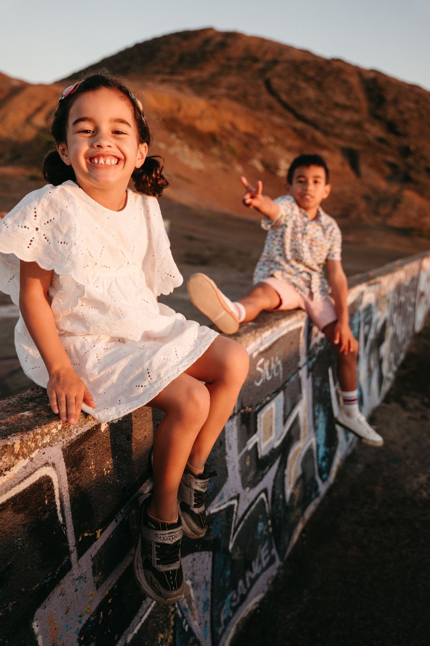A smiling girl in a white dress sits on a graffiti wall with a boy at sunset.