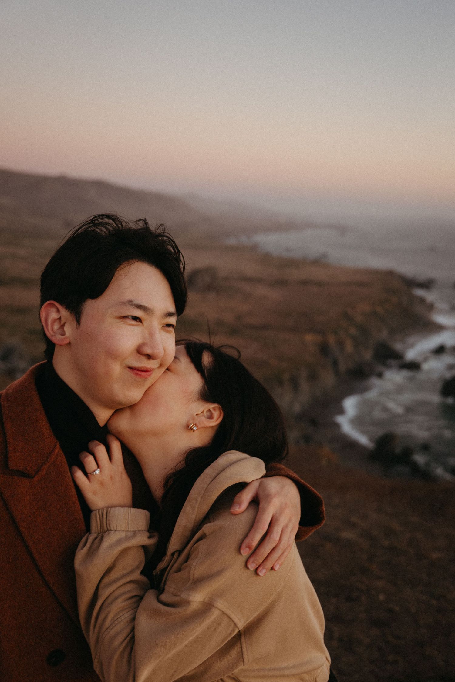 A woman kisses a smiling man on the cheek as they embrace on a coastal bluff overlooking the ocean at a soft pink sunset.