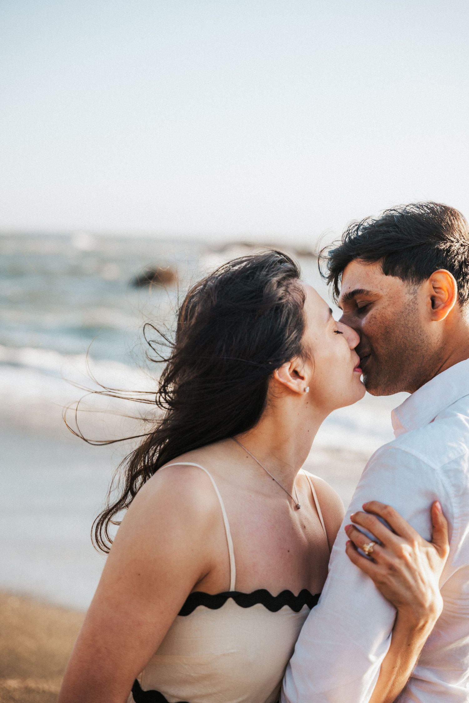 Romantic couple sharing a kiss on a windy beach, woman in white dress with black trim embracing man in white shirt.