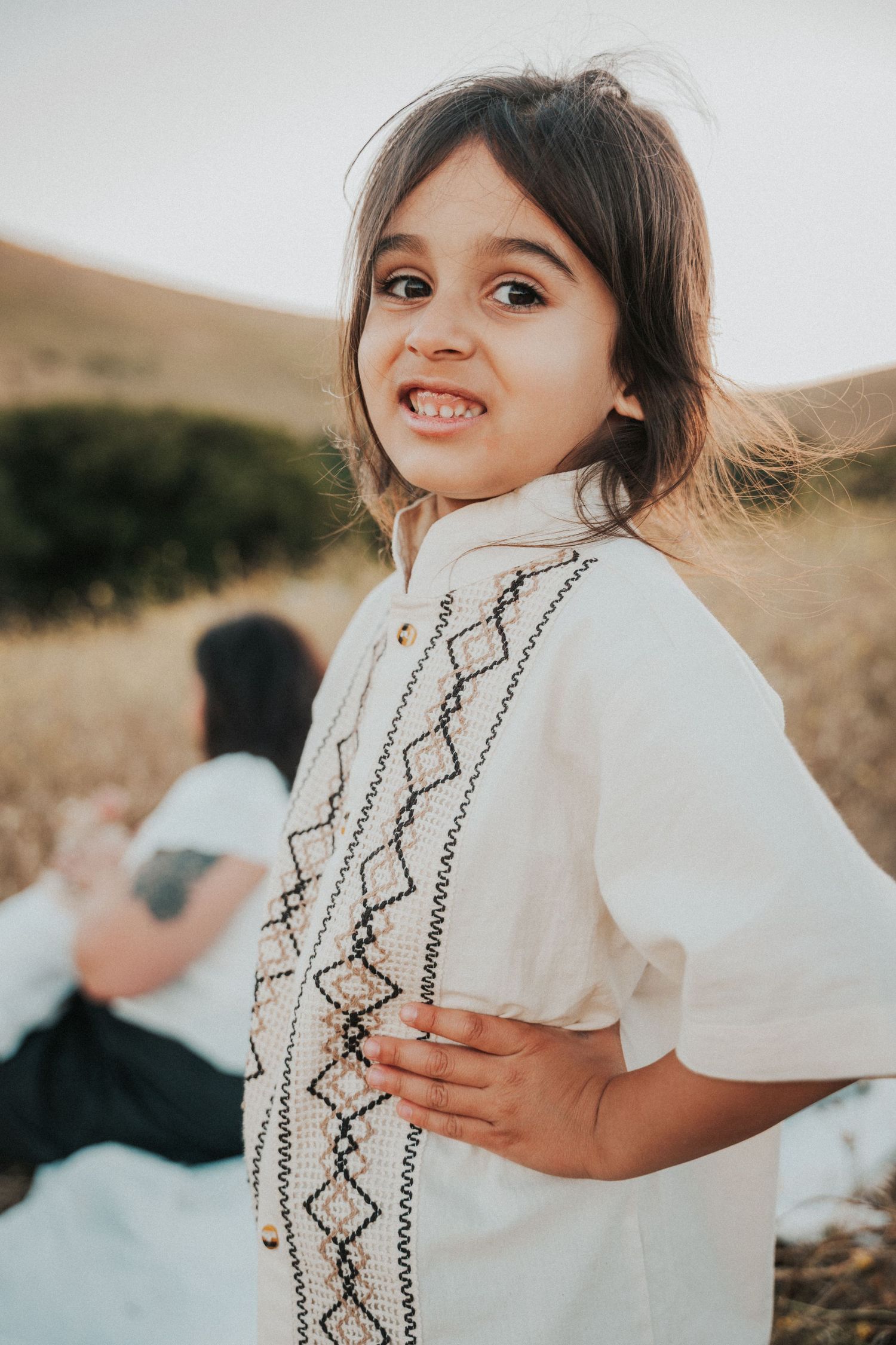 A smiling young girl in a white embroidered dress poses outdoors in a sunny field with hills in the background.
