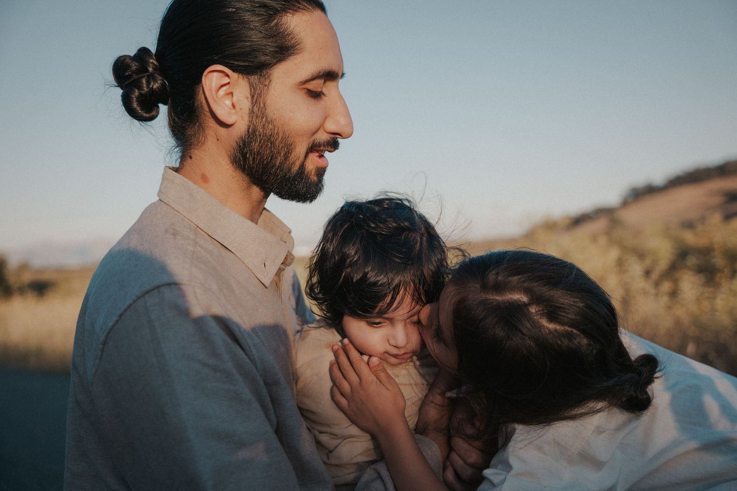 A bearded man with a bun holds a young child as a woman leans in to kiss the baby outdoors at sunset.