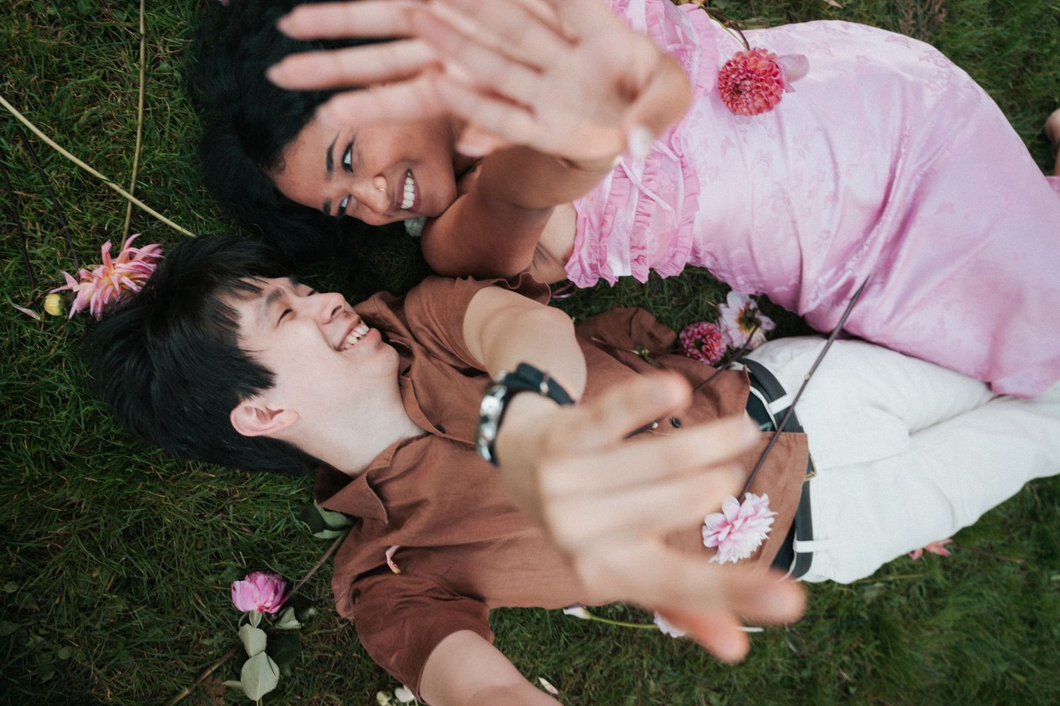 Couple lying on grass laughing, surrounded by pink flowers, reaching hands toward camera in joyful moment.
