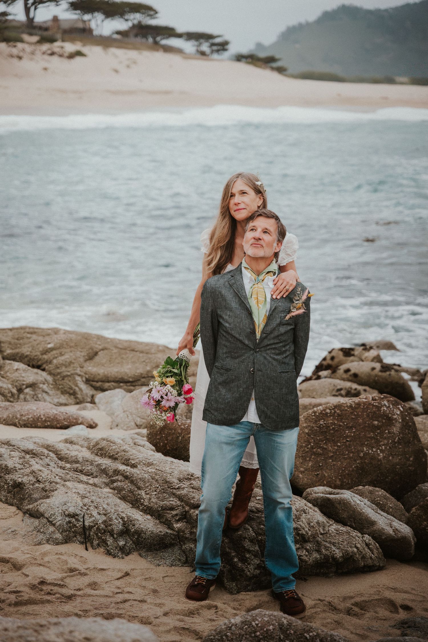 Couple embracing on rocky beach, woman holding colorful bouquet, ocean waves in background.