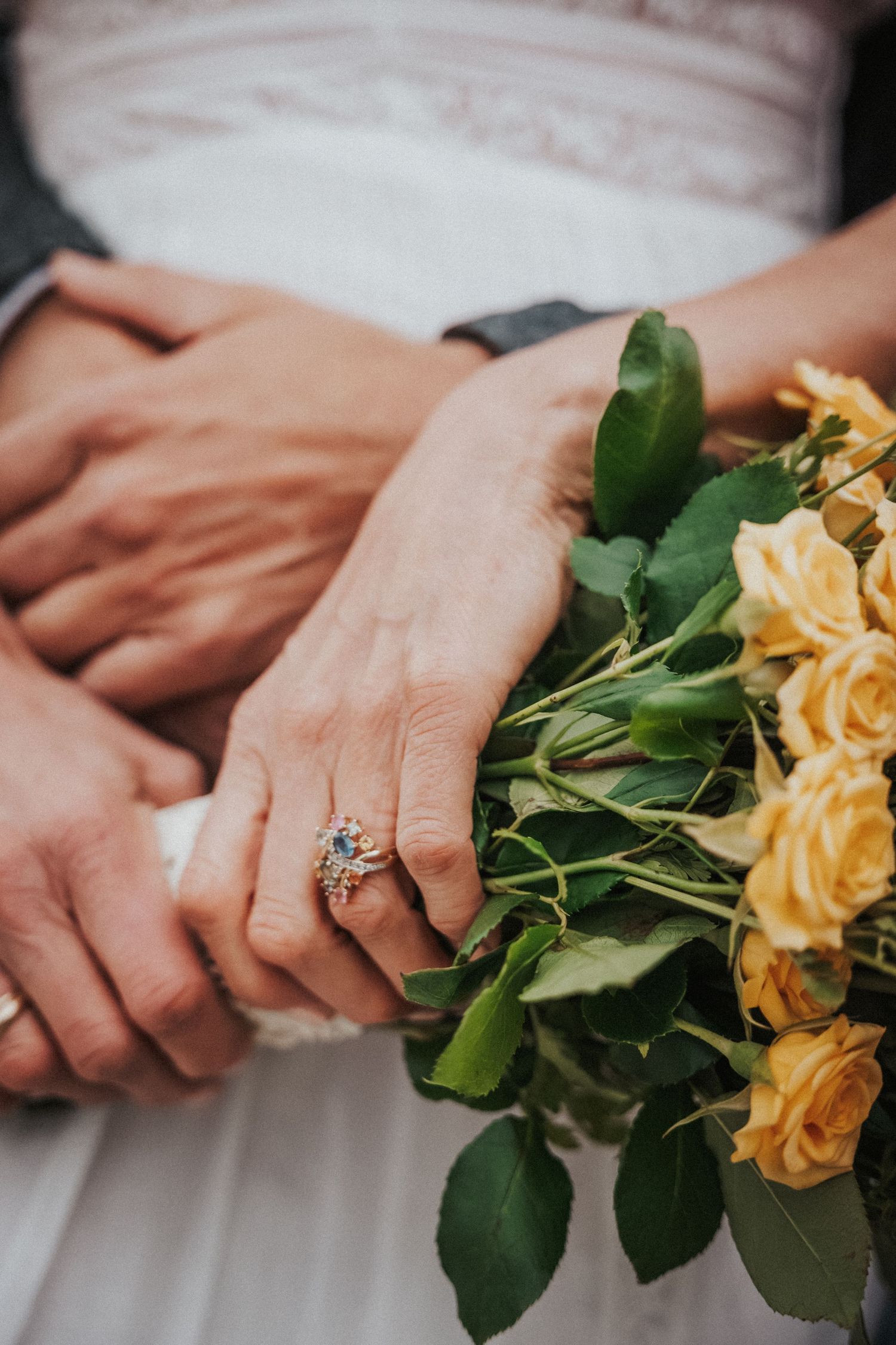 Bride and groom holding hands with yellow rose bouquet, featuring a colorful gemstone engagement ring on wedding day.