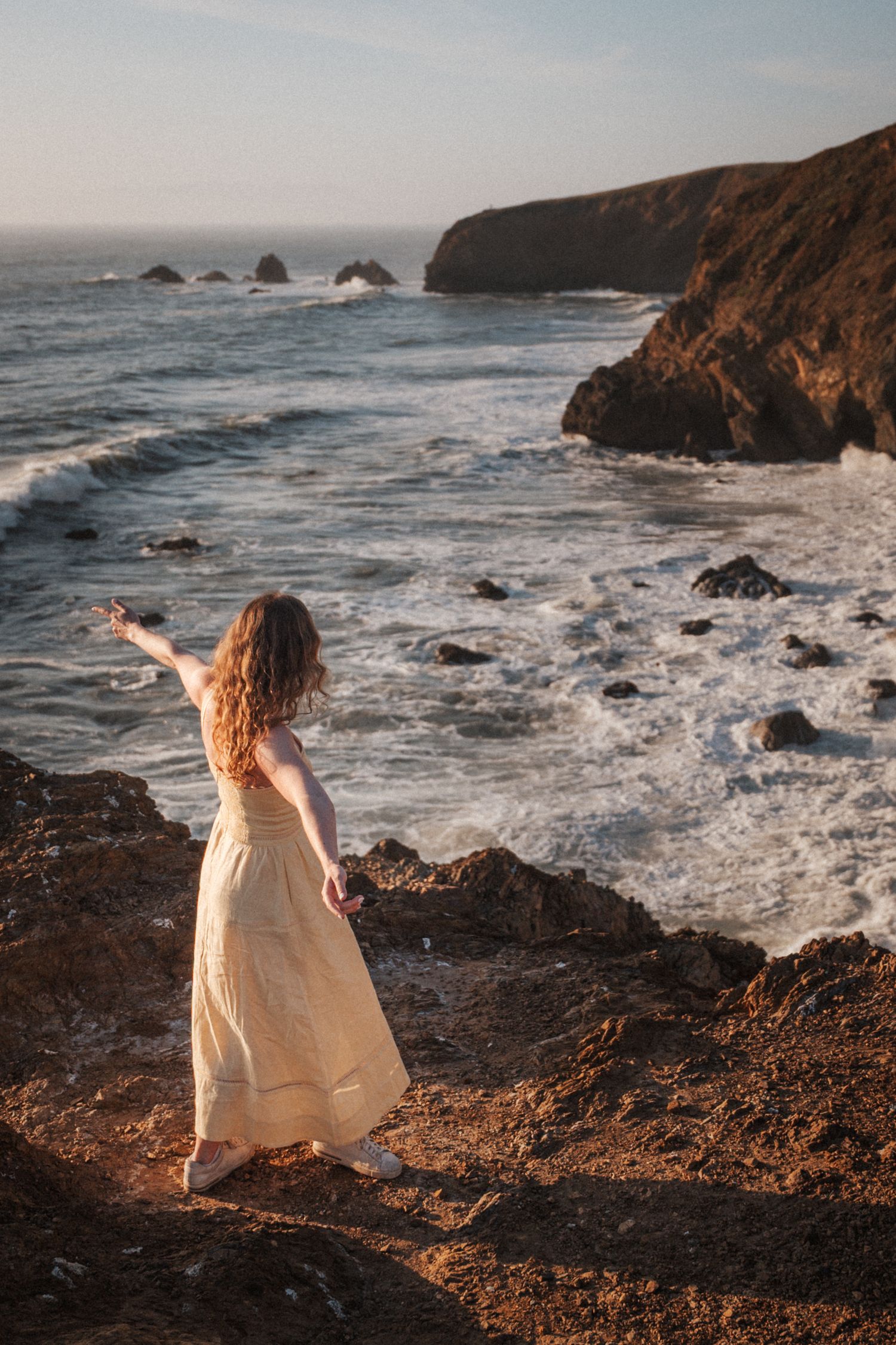 Woman in cream dress standing on rocky coastal cliffs overlooking dramatic ocean waves at sunset.