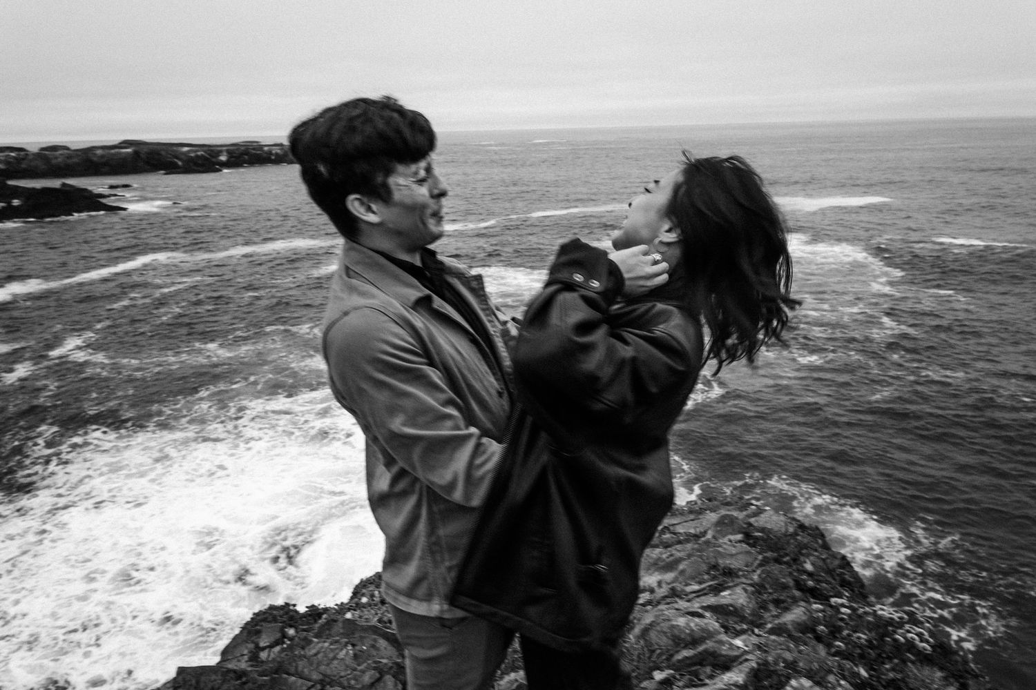 Black and white photo of a couple embracing on rocky coastal cliffs with ocean waves in the background.