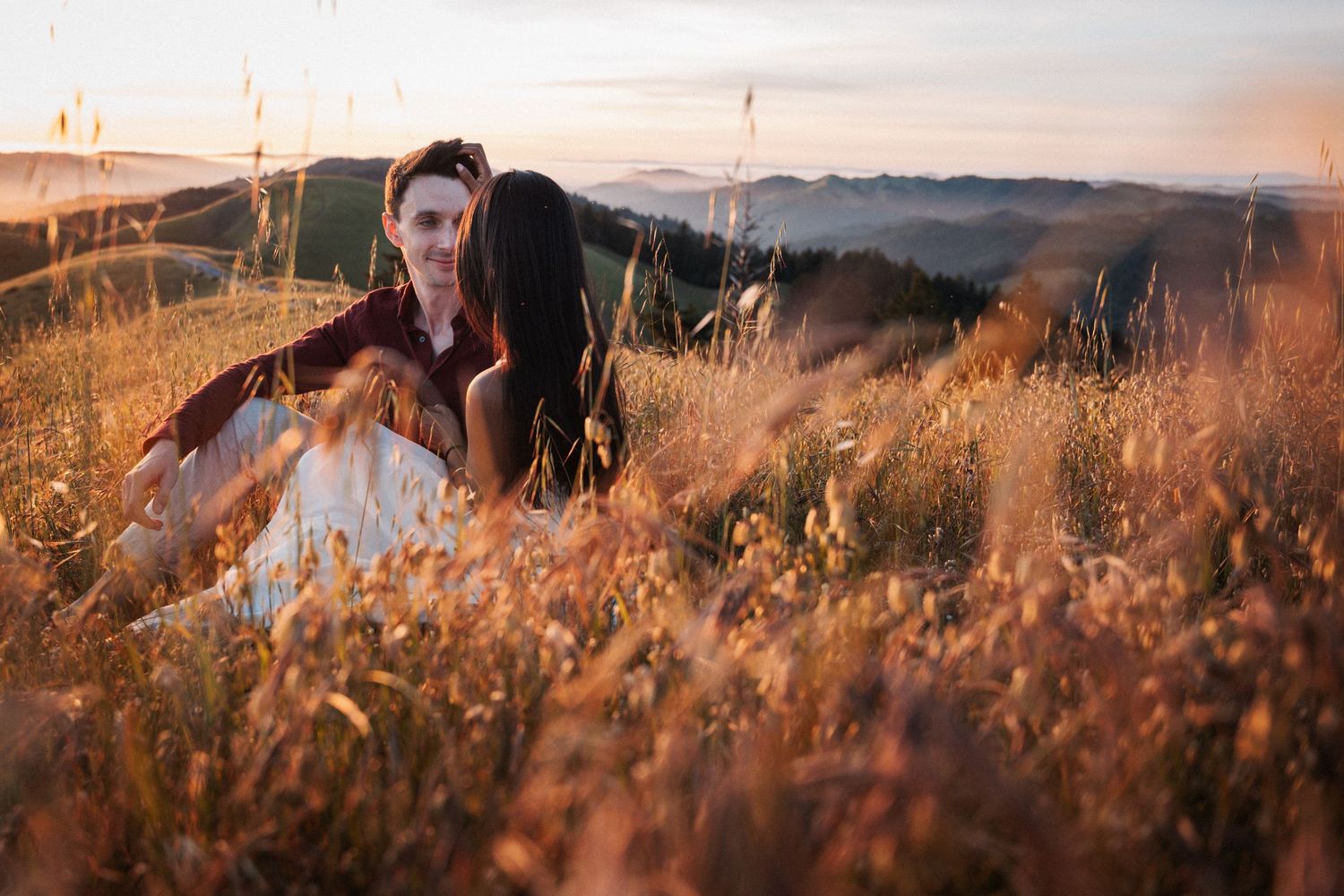 Romantic couple embracing in golden tall grass field with rolling hills and sunset in background.