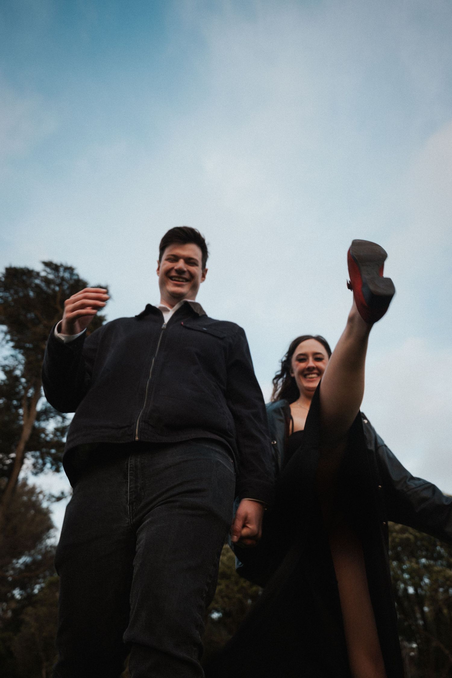 Joyful couple in black outfits laughing and kicking outdoors against a cloudy sky.