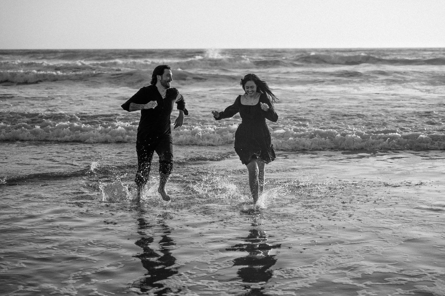 Black and white photo of a couple running and splashing through ocean waves on a beach.