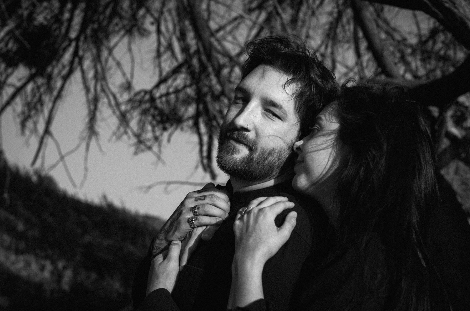 Black and white romantic portrait of bearded man and woman embracing under bare tree branches.