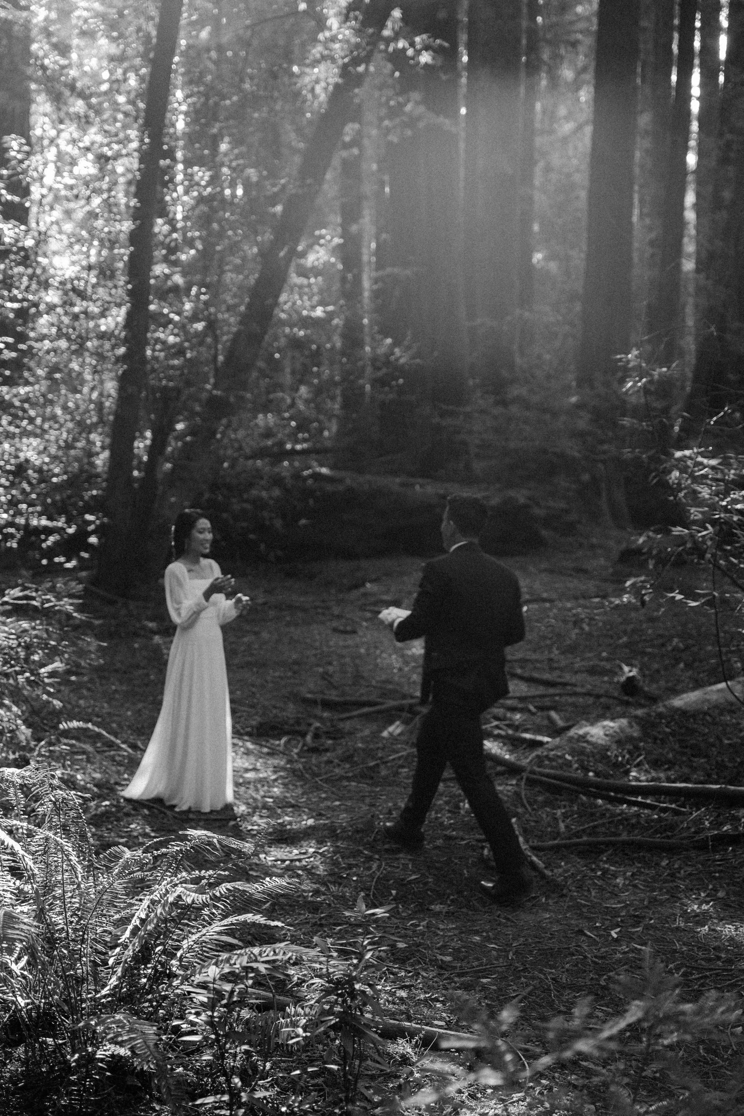 Black and white photo of a bride and groom exchanging vows in a sunlit forest clearing.
