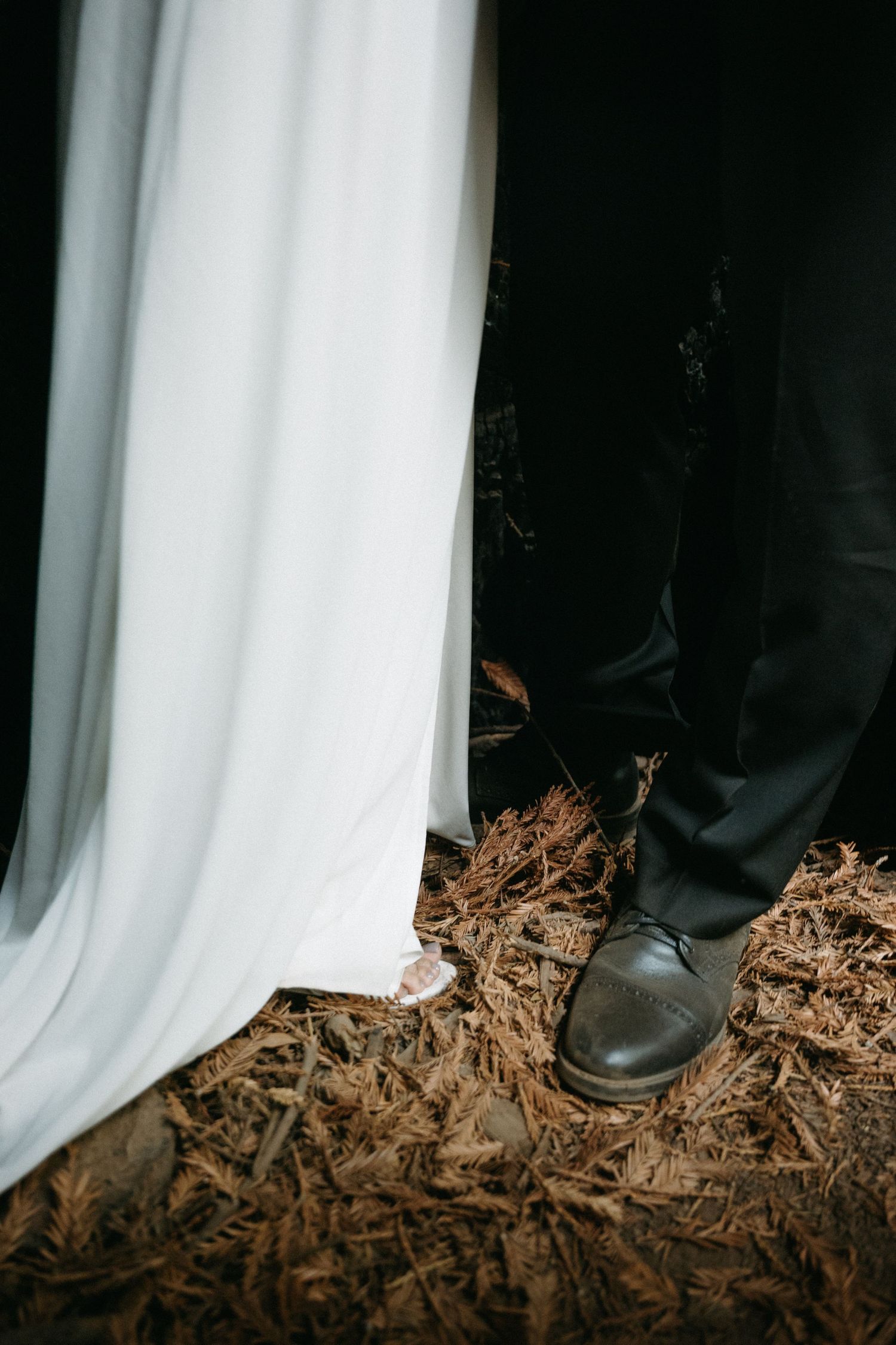 Close-up of a bride's white wedding dress hem and groom's black dress shoes standing on pine needle covered ground.