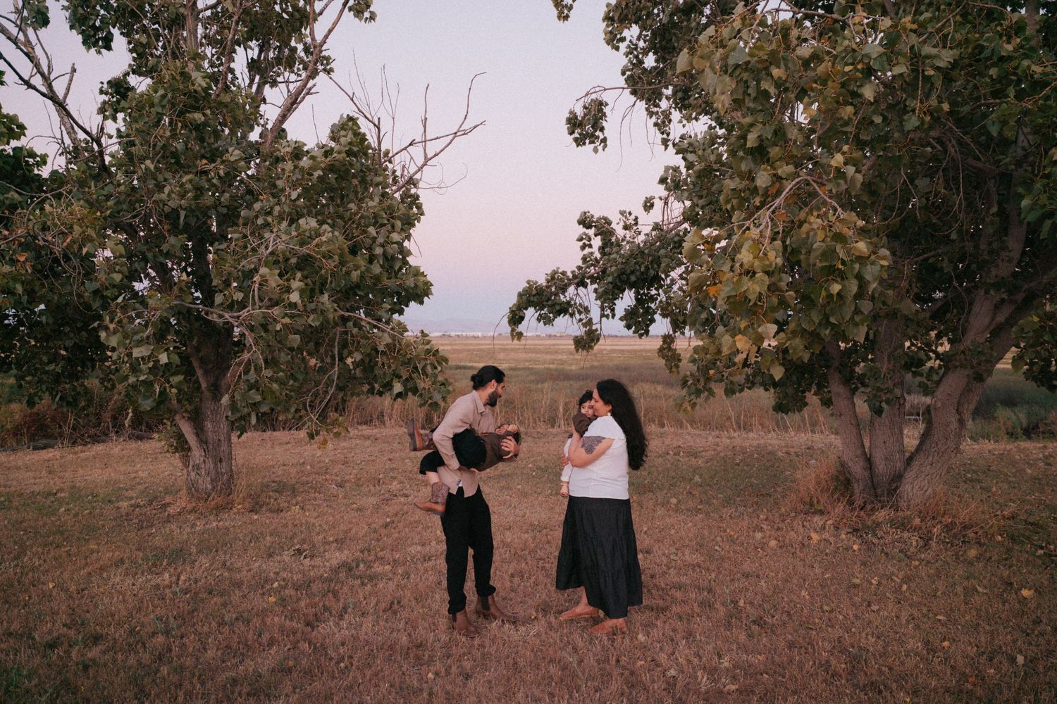 A family of three stands among sparse trees on dry grassland at dusk, with a child being held by one adult.