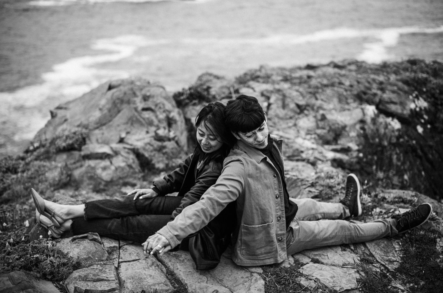 Black and white photo of couple sitting back to back on coastal rocks by the sea.