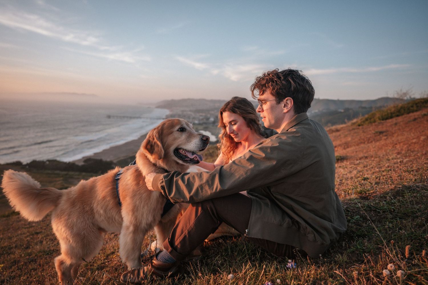 Couple sitting on hillside at sunset with their golden retriever dog.