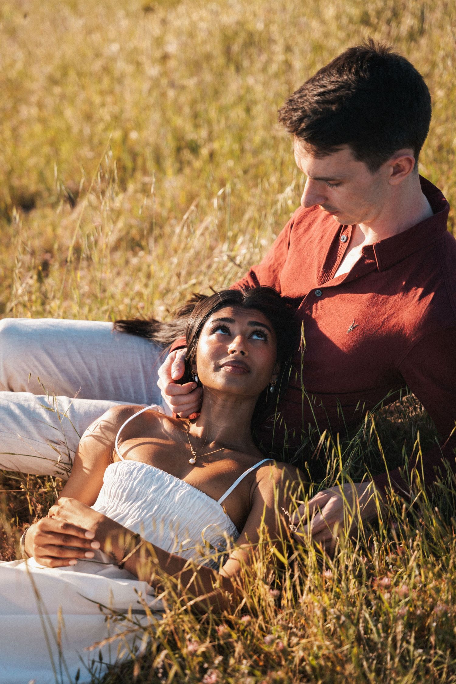 Romantic couple relaxing in golden grass field, woman in white dress lying on man's lap gazing up at him.