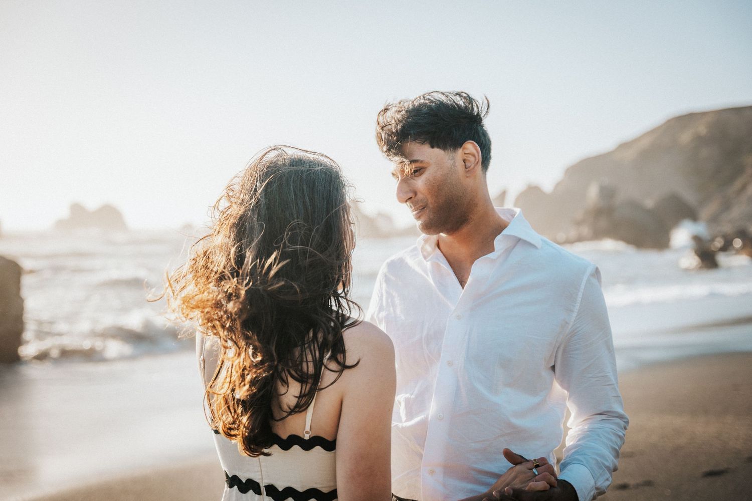 Couple sharing a romantic moment on a sunny beach with ocean waves and rocky cliffs in the background.