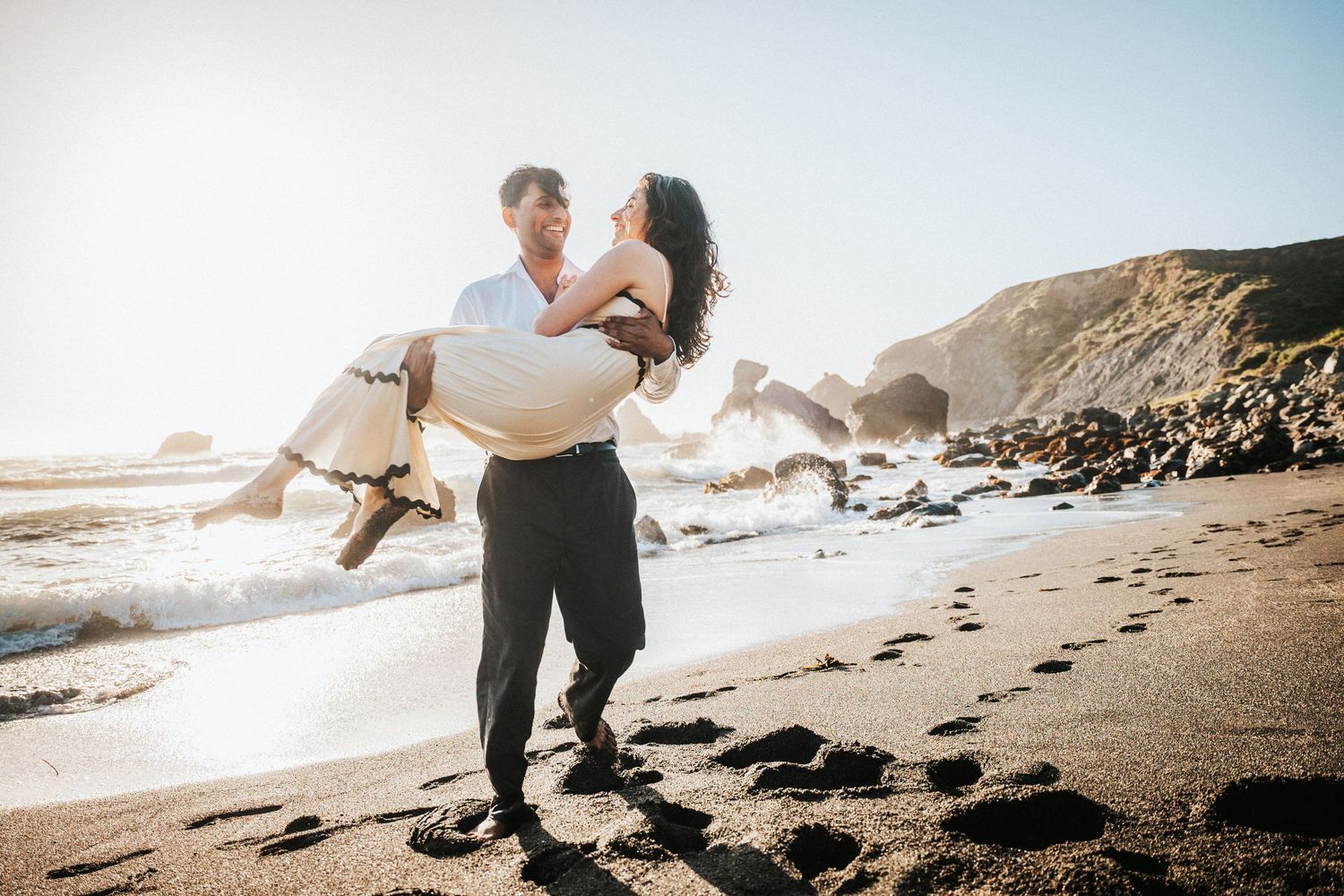 Man carrying woman in his arms on sunny beach, laughing couple in romantic beach engagement photography.
