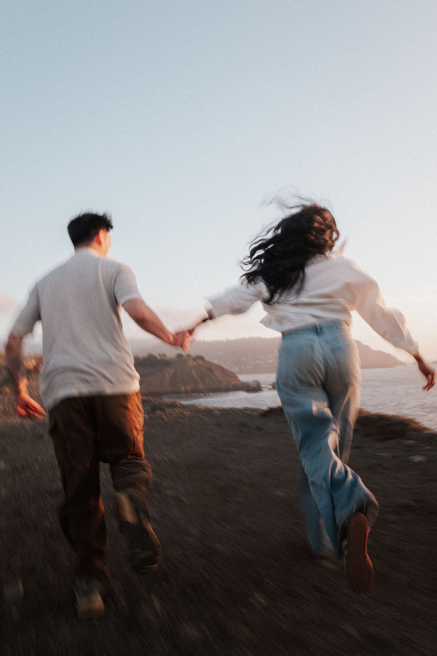 Blurred motion shot of couple holding hands running along coastal cliffside at golden hour sunset.