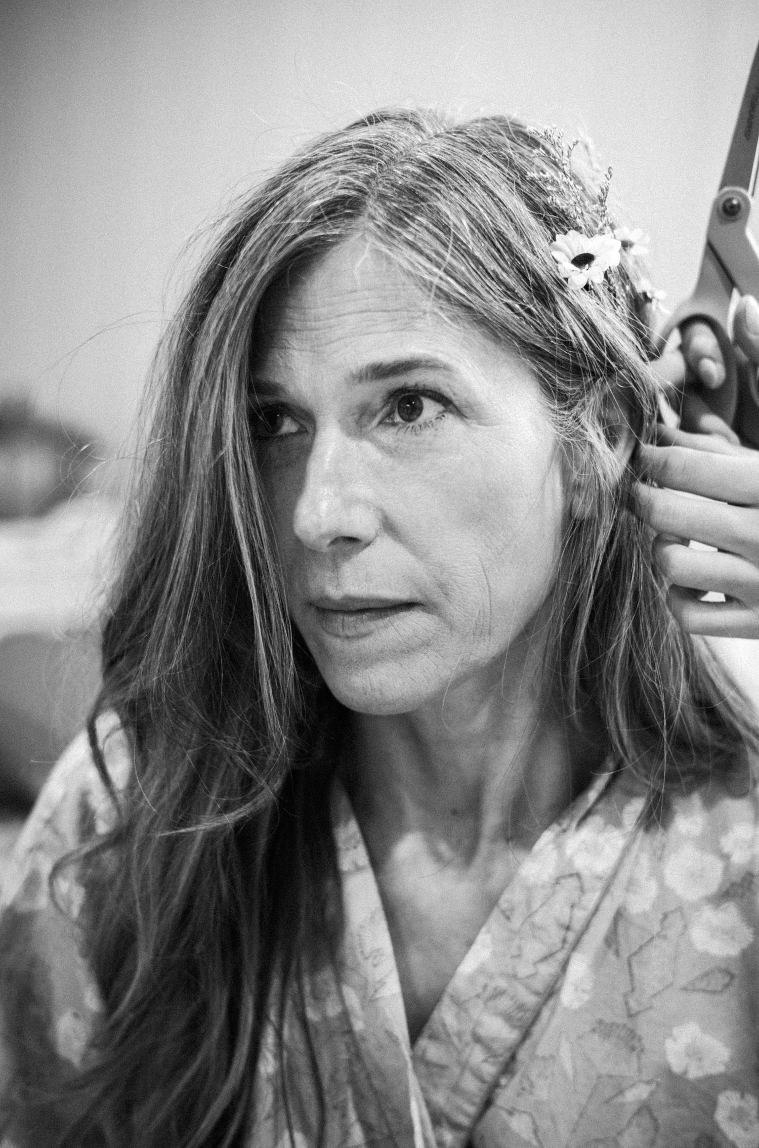 Black and white portrait of a middle-aged woman with long hair and flowers, holding scissors near her head.