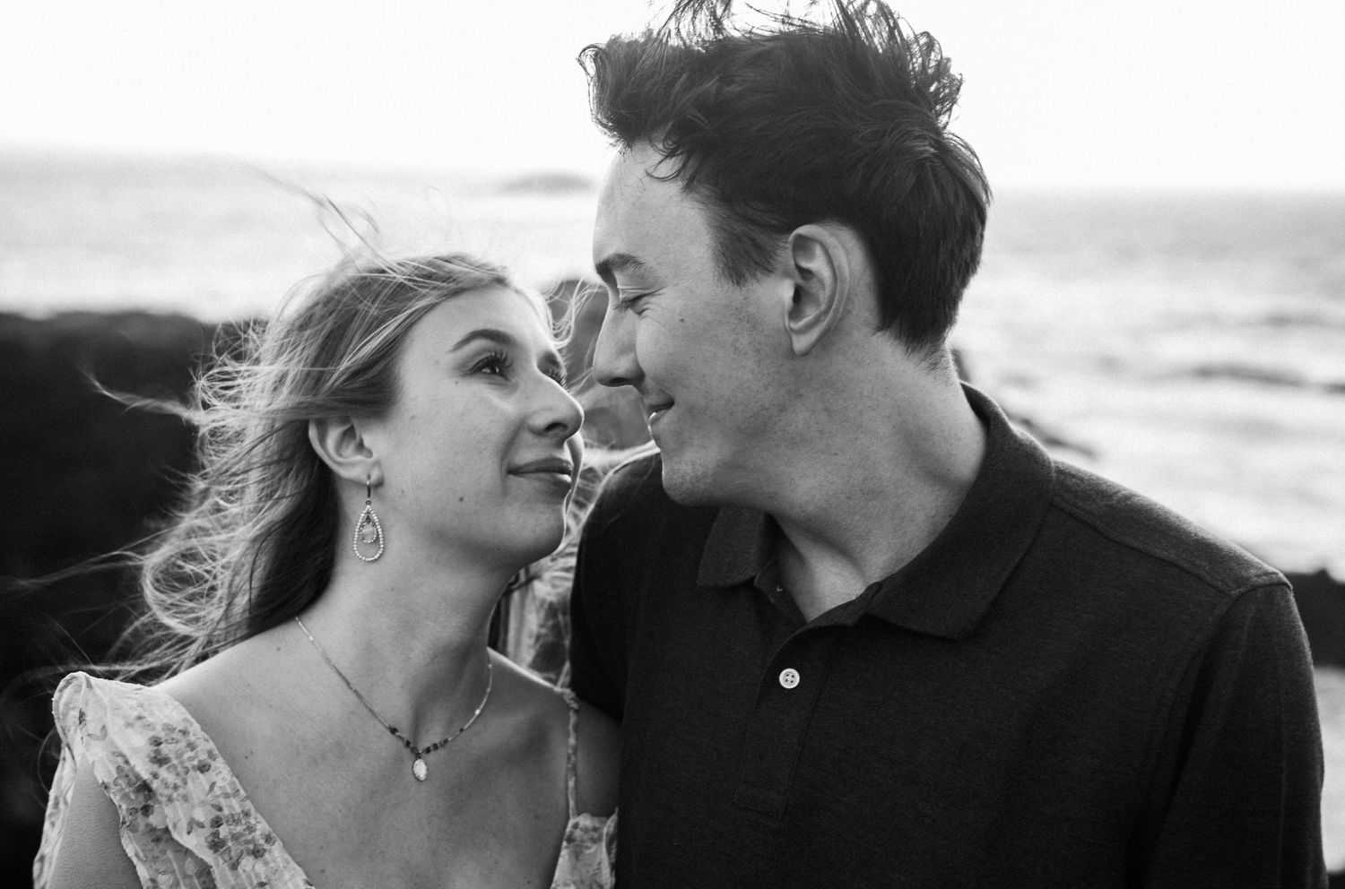 Black and white photo of a smiling couple sharing a romantic moment by the ocean, woman wearing floral dress.