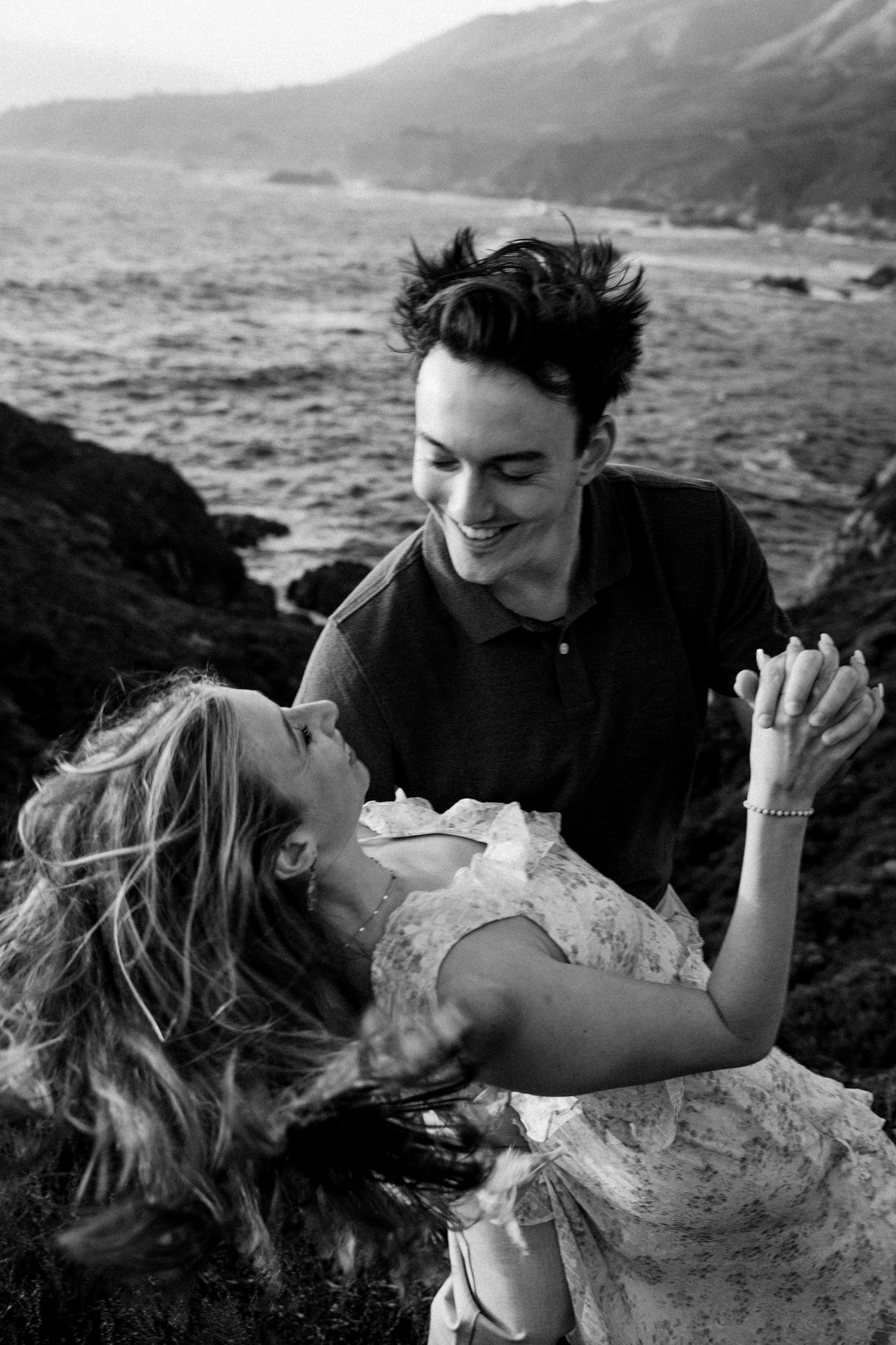 Black and white photo of a laughing couple embracing on rocky coastal cliffs with ocean waves in the background.