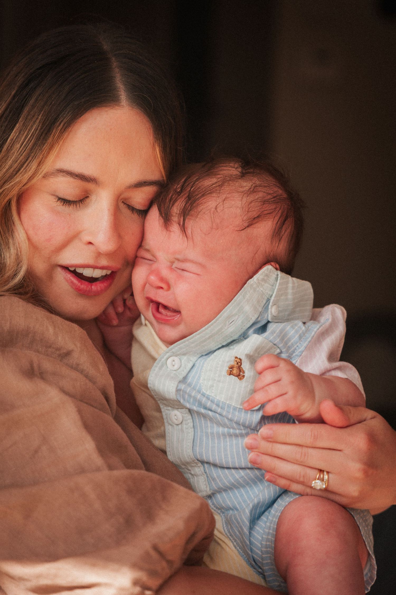 Mother tenderly holding her crying newborn baby dressed in a light blue outfit indoors.