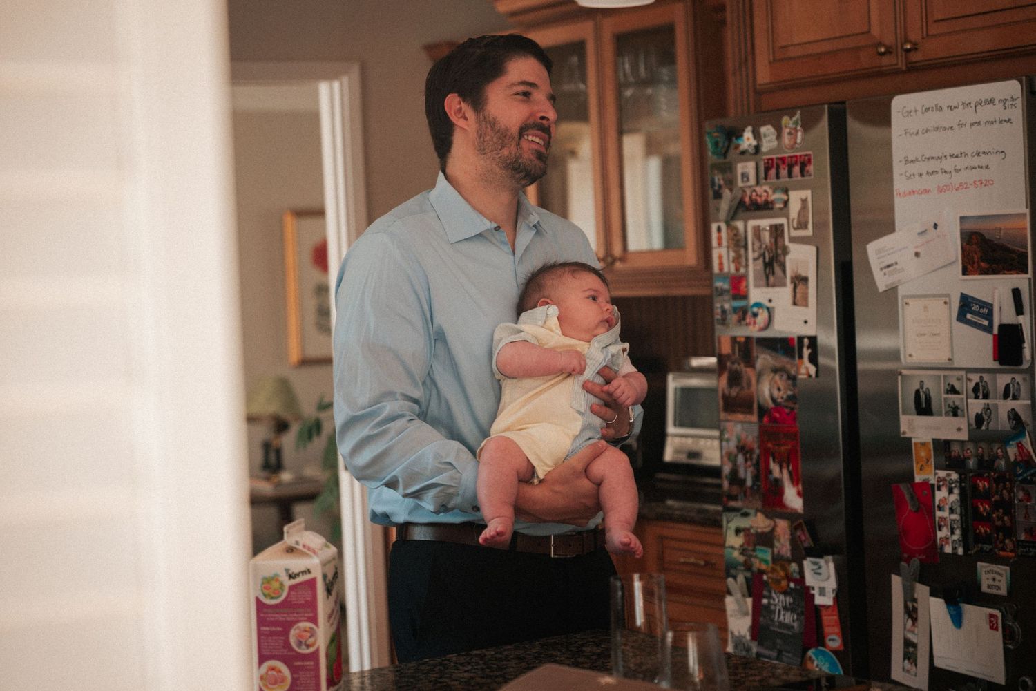 A smiling father in a blue shirt holds his newborn baby in a cozy kitchen with a refrigerator covered in notes.