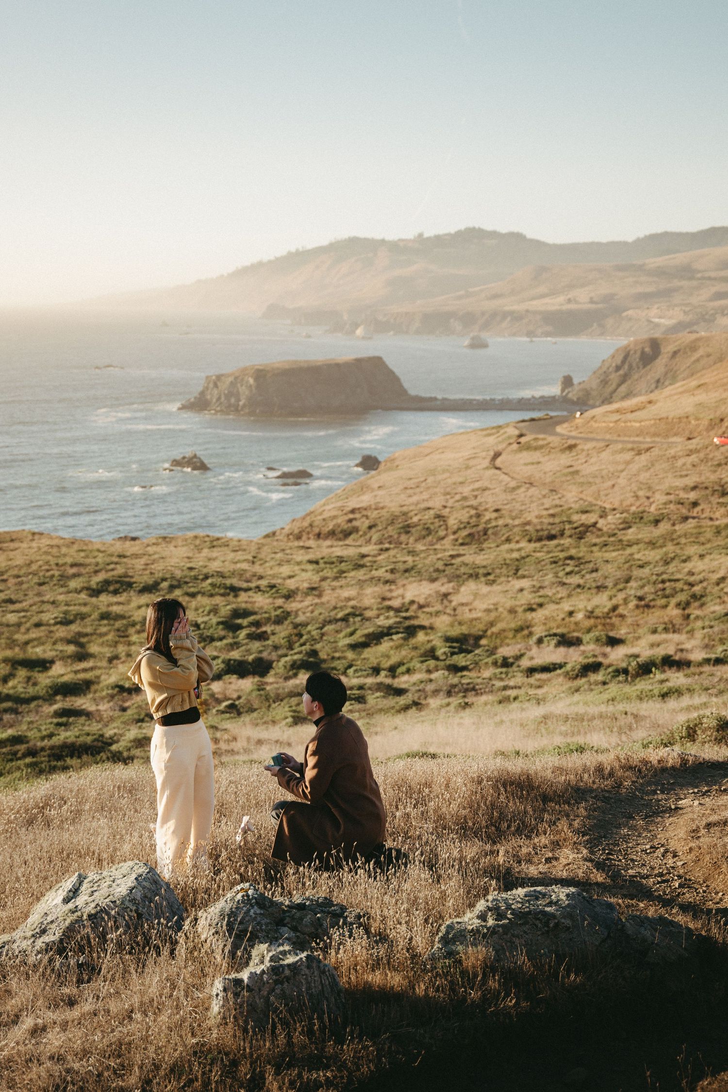 A man kneels with a ring box as a woman covers her face in surprise during a coastal cliff proposal at golden hour.