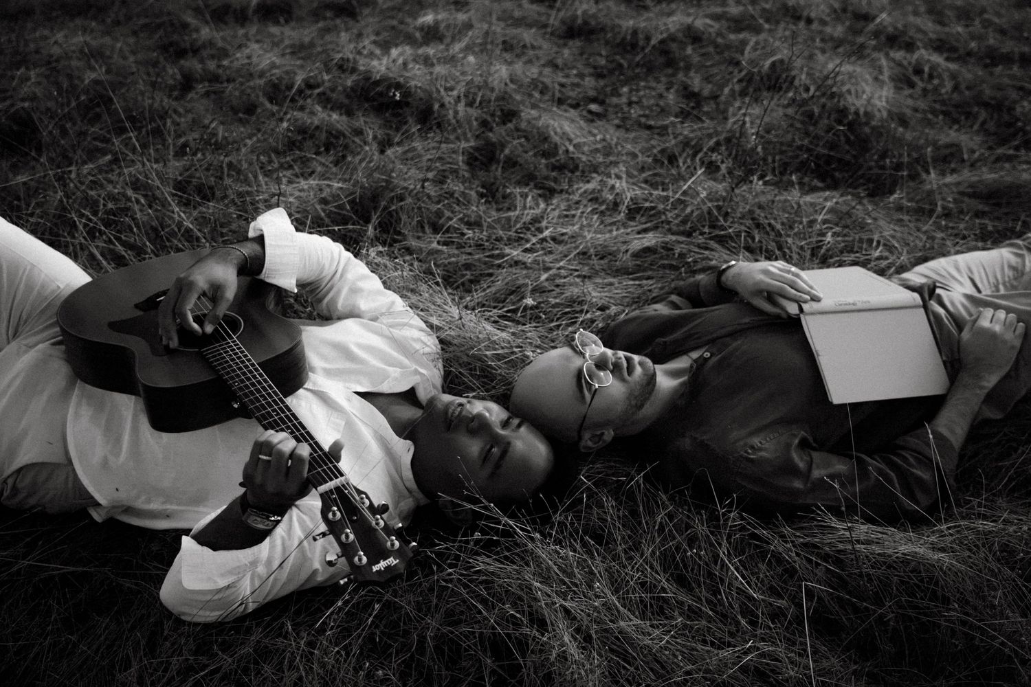 Black and white photo of two people lying in grass, one playing acoustic guitar while the other reads a book.