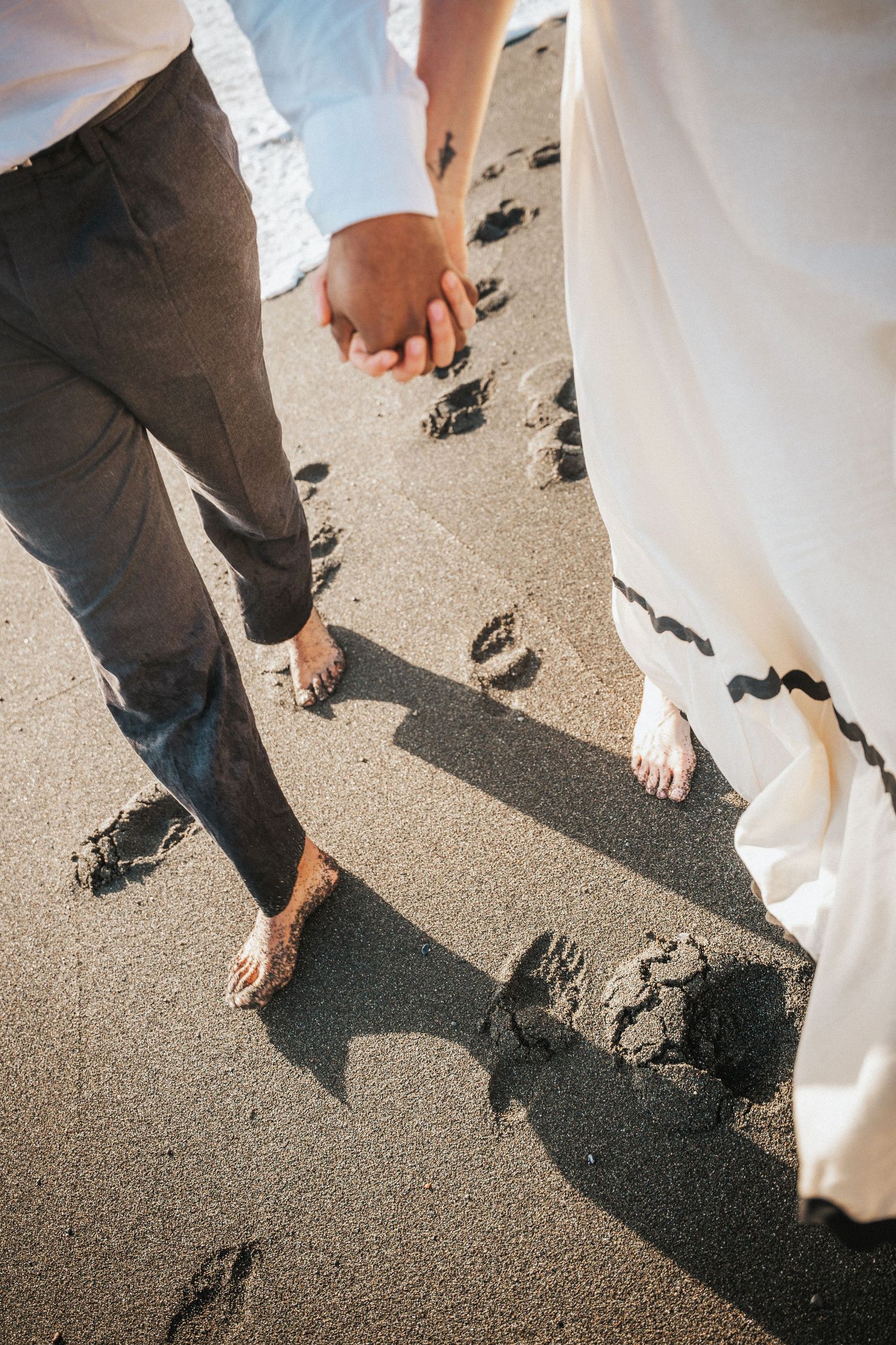 Barefoot couple holding hands walking on dark sand beach, leaving footprints, wearing wedding attire.Photography by The Y.K. Studio.