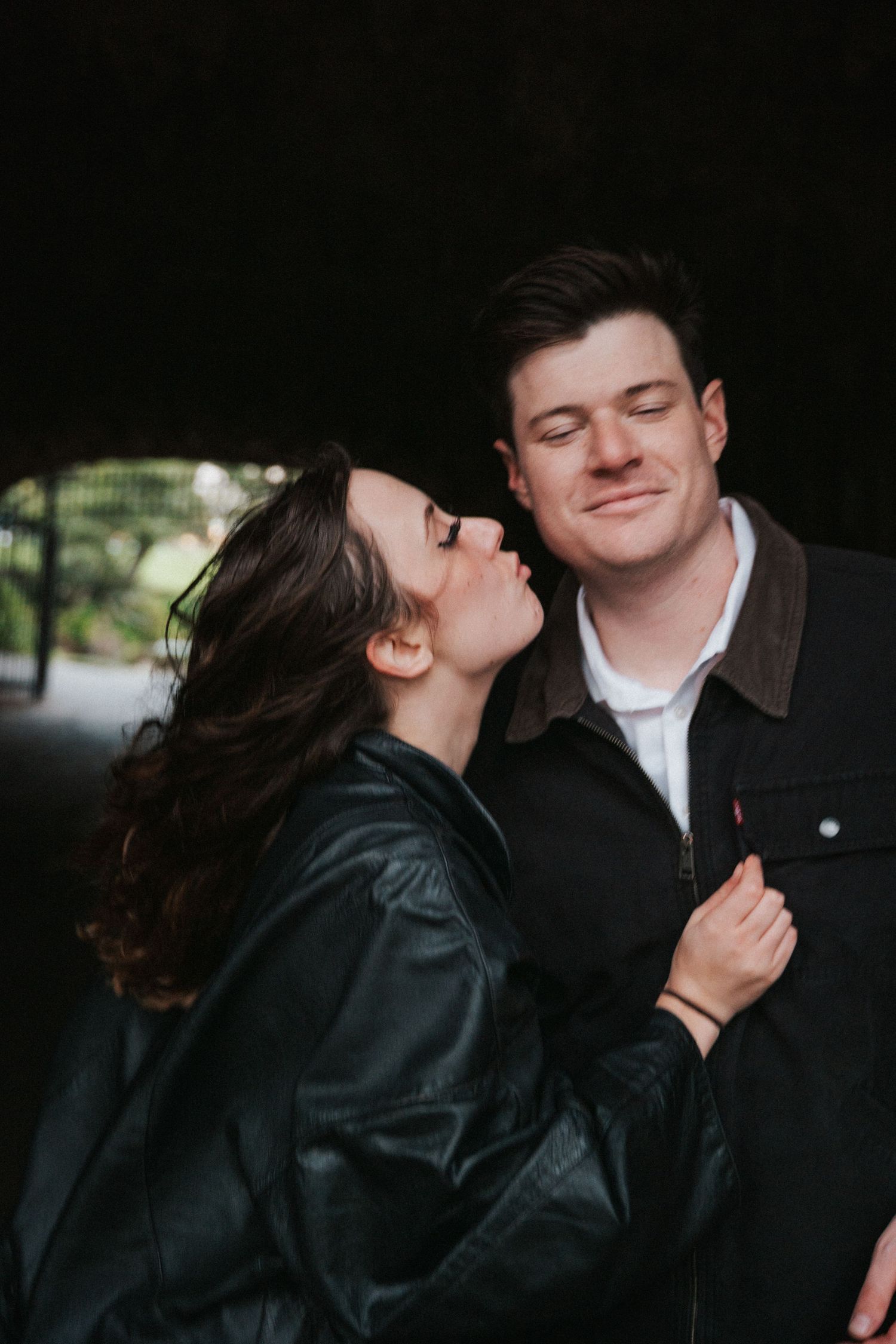 A couple shares a tender moment under a bridge, woman in leather jacket leaning in to kiss smiling man in Levi's jacket.Photography by The Y