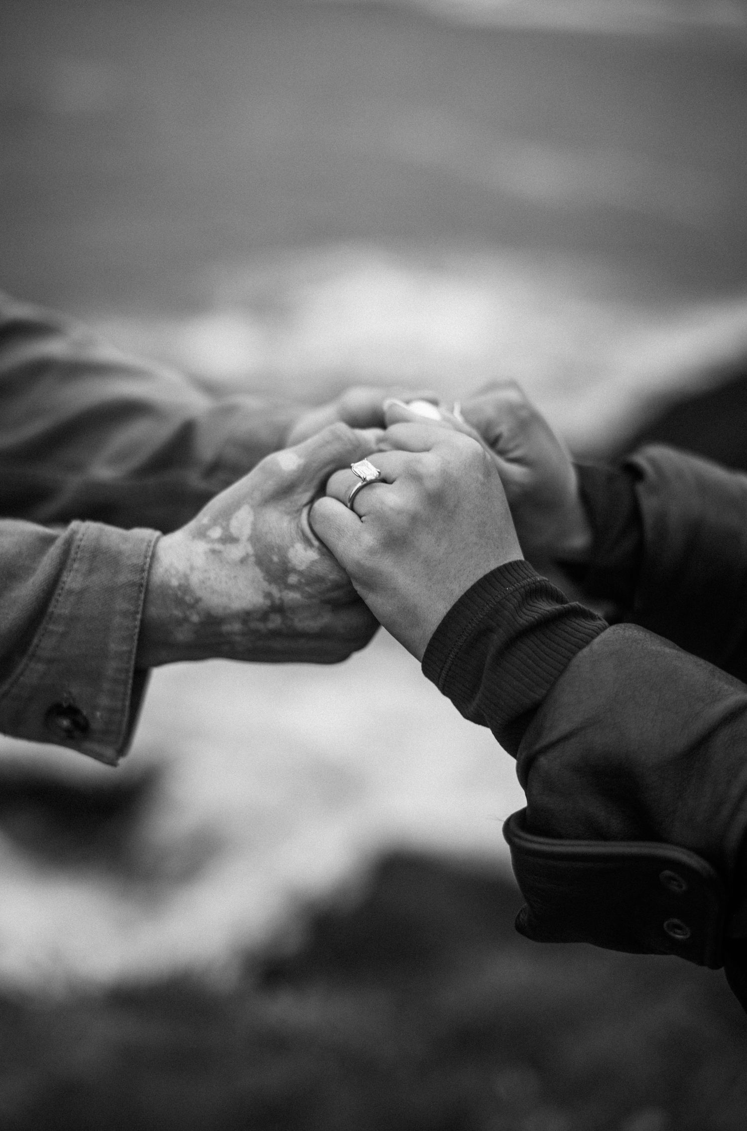 Black and white photo of a couple holding hands, showcasing an engagement ring, with a blurred ocean background.