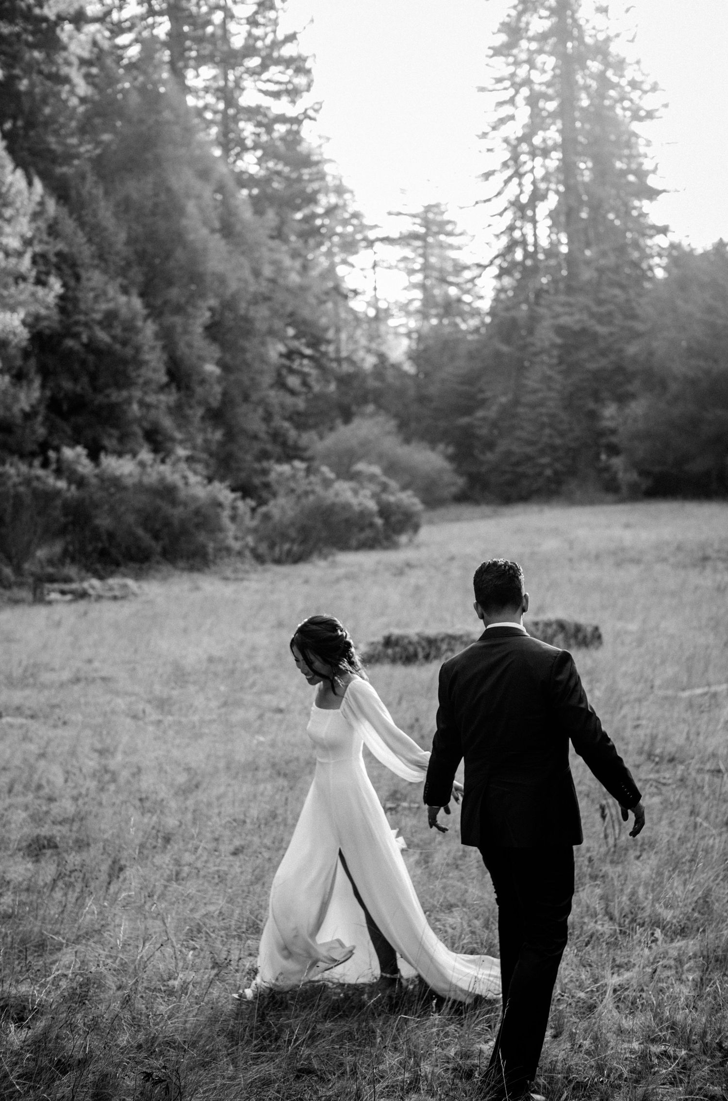 Black and white photo of a bride and groom walking hand in hand through a meadow surrounded by tall trees.