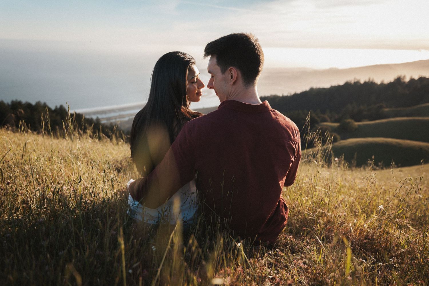 Couple sitting together in golden meadow grass sharing a romantic moment with scenic coastal hills at sunset.