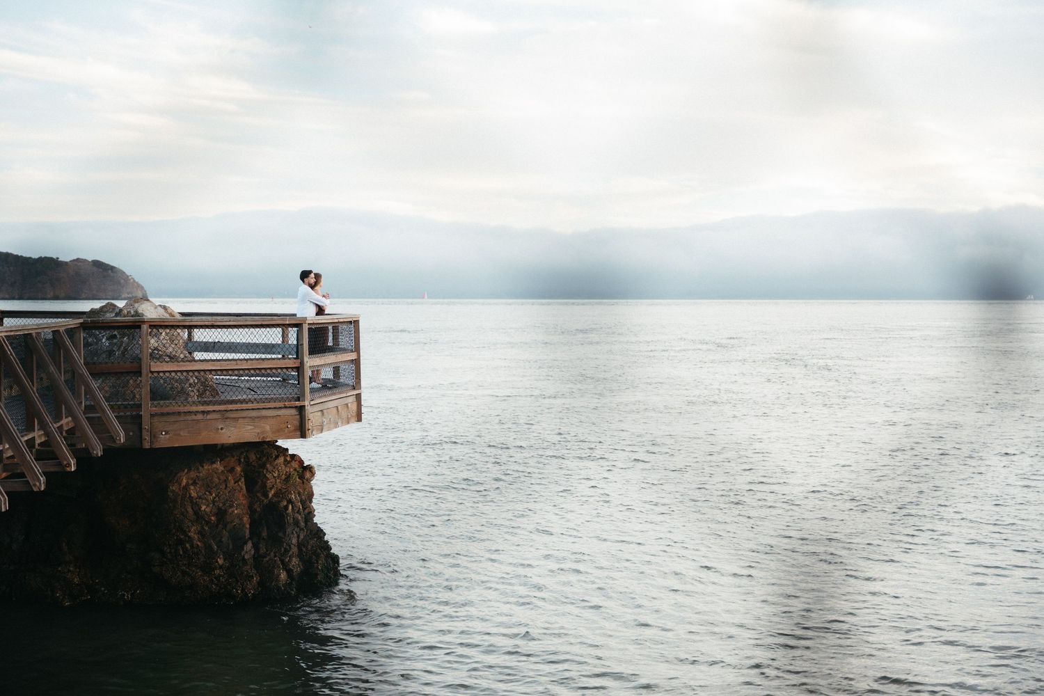 Couple embracing on a wooden cliffside deck overlooking calm ocean waters with misty mountains in the background.