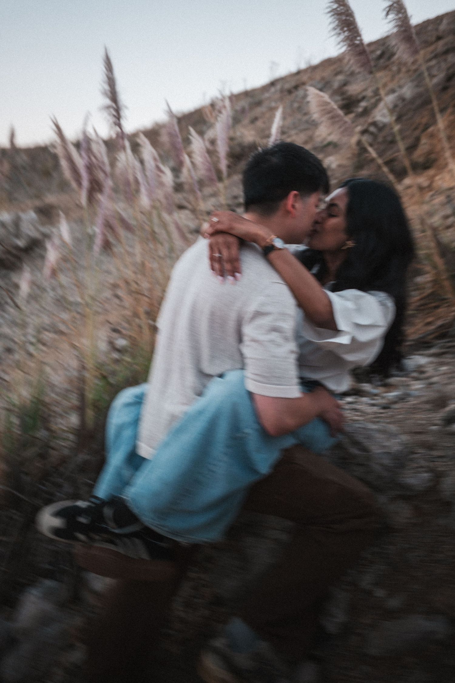A couple shares a romantic kiss outdoors, surrounded by wild grasses and rocky terrain in soft evening light.