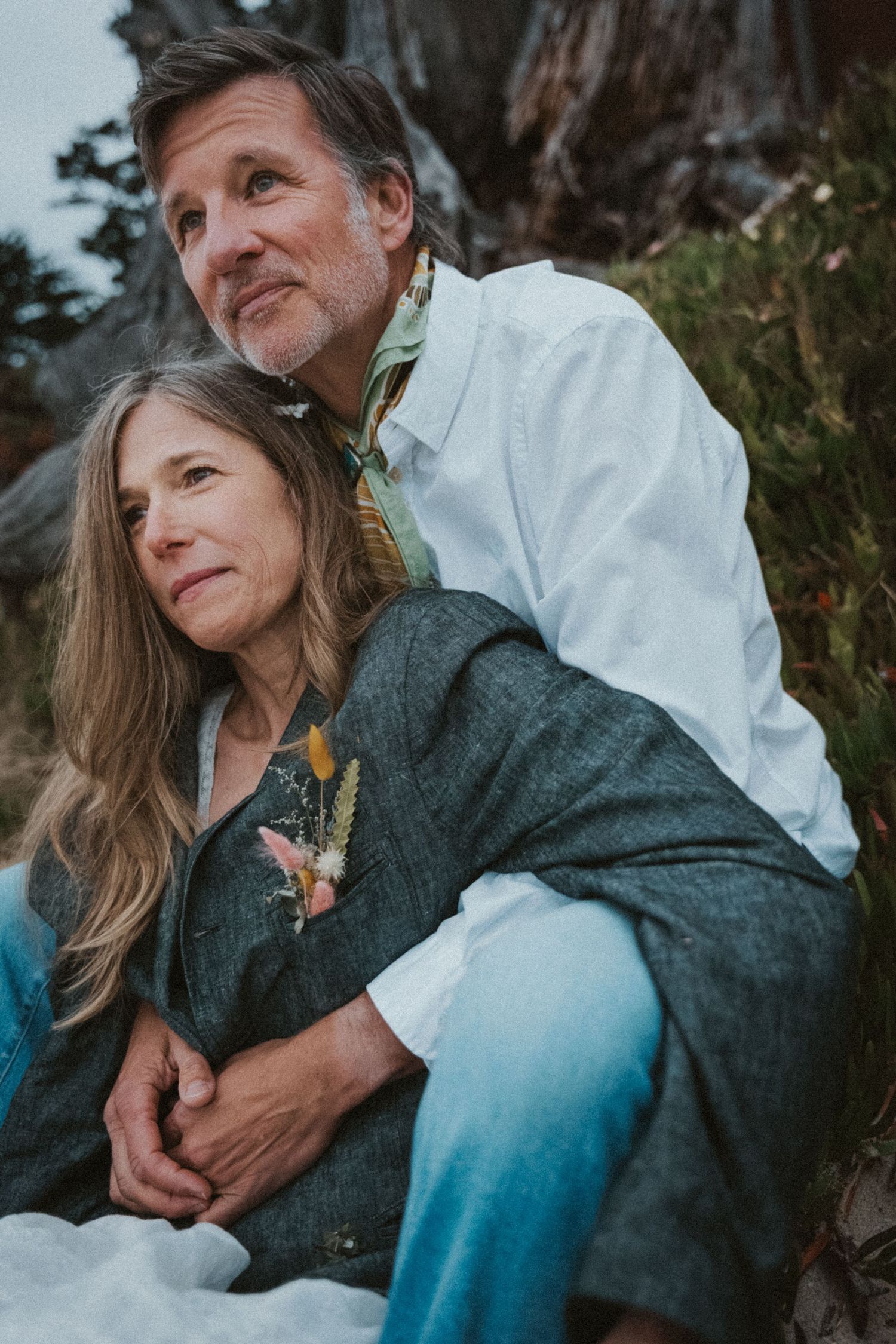Couple sitting together outdoors near a tree, embracing and gazing into the distance peacefully.