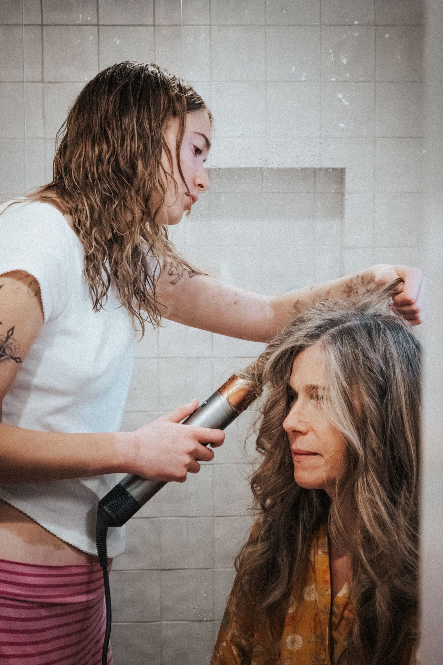 Young woman with wet hair uses a curling iron to style an older womans long wavy brown hair in a tiled bathroom.