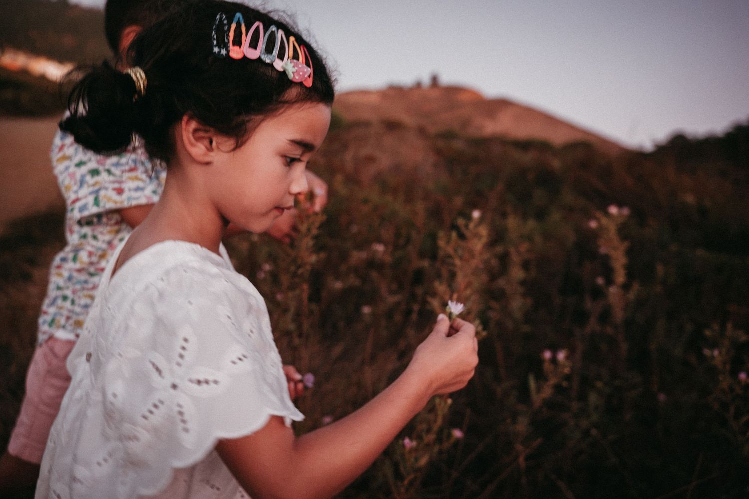 Young girl in white dress examining a wildflower in a field at dusk wearing a tiara headband.