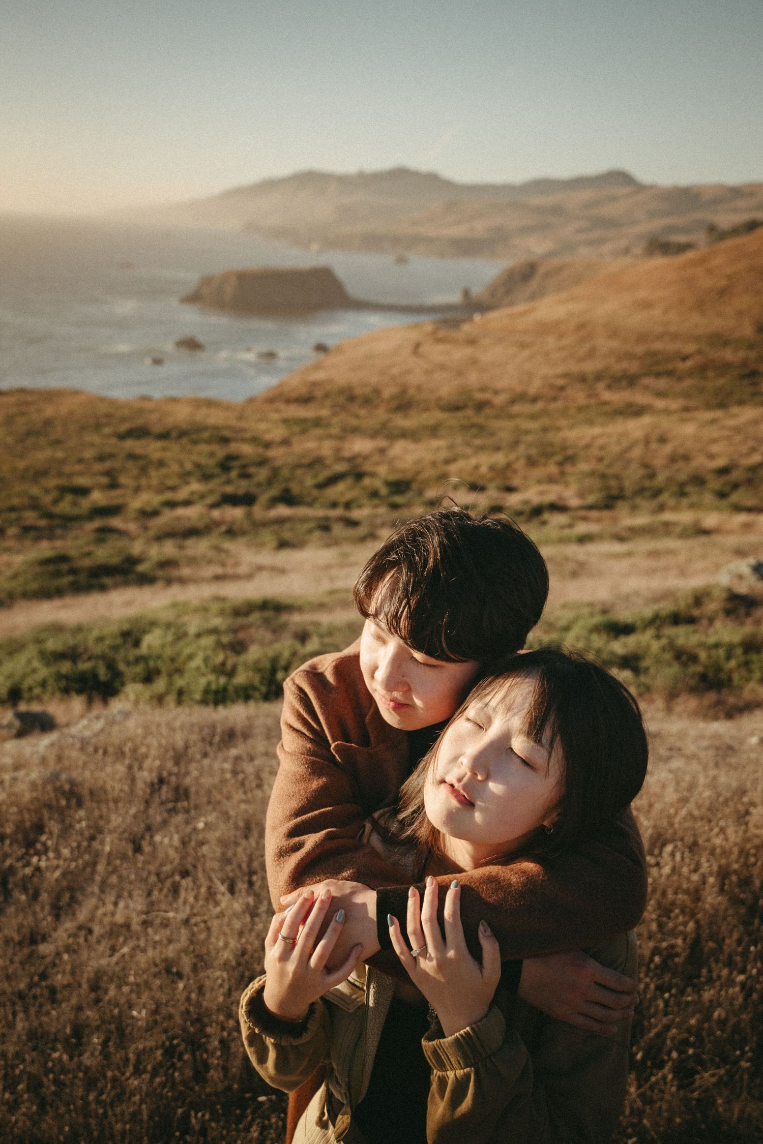 Two people embrace in golden light on a grassy coastal hillside, with ocean and rolling hills stretching behind them.