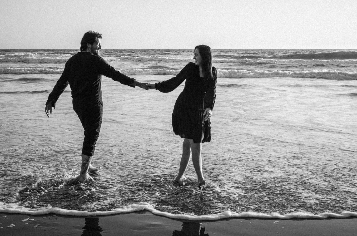 Black and white photo of a couple holding hands and wading in ocean waves on a beach.