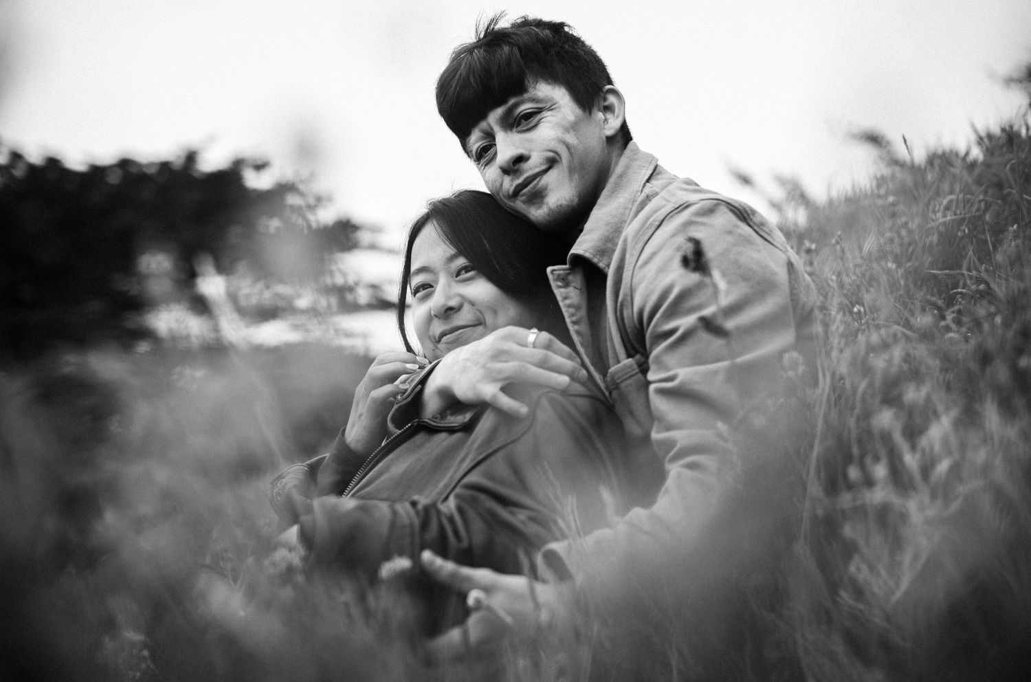 Black and white photo of smiling couple embracing warmly in tall grass field.