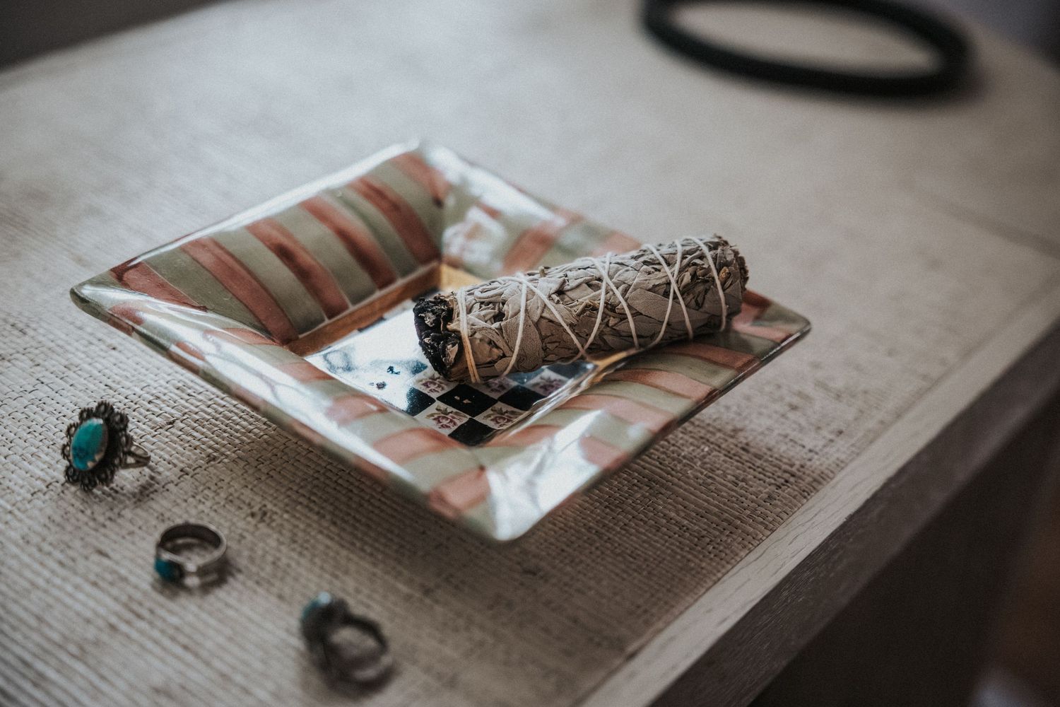 A decorative ceramic dish holding a sage smudge stick, surrounded by turquoise and silver rings on a wooden table.