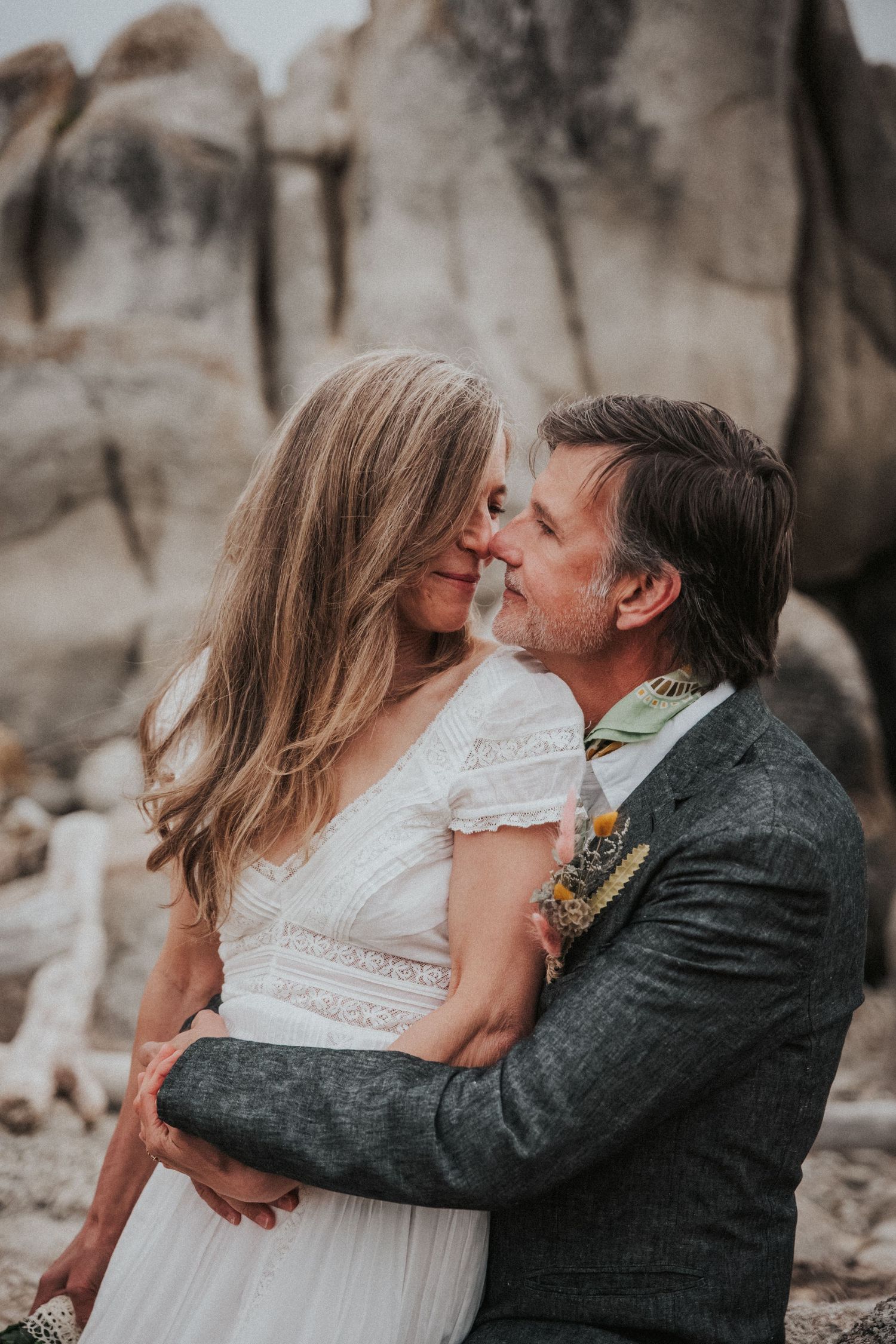 Couple in wedding attire embracing and touching foreheads among rocky boulders outdoors.