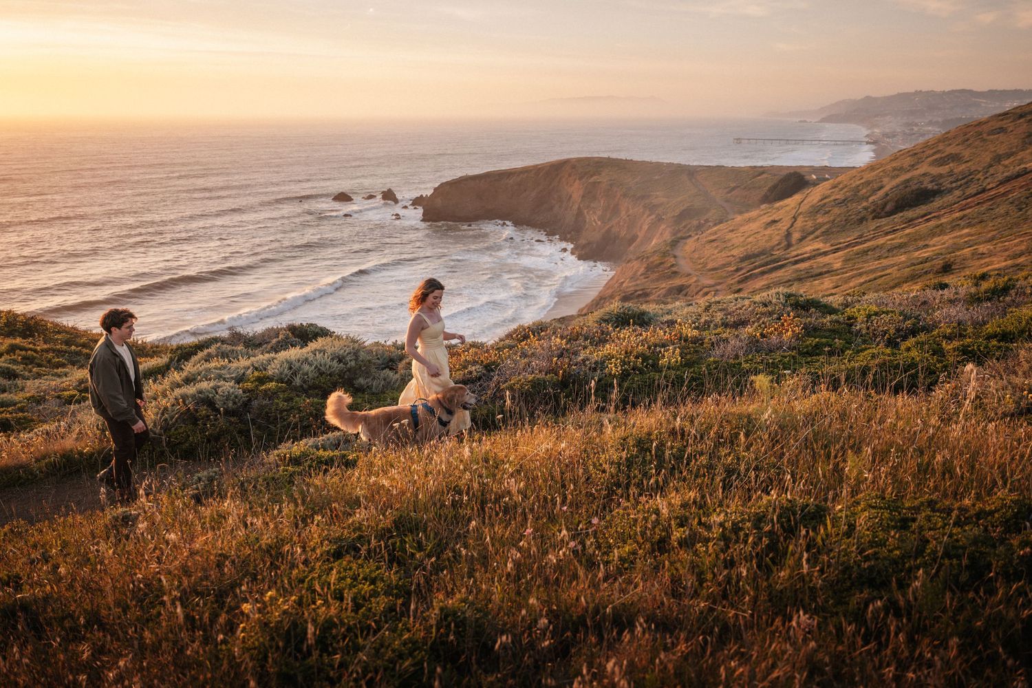 Couple with golden dog on coastal cliffside at sunset, golden hour light over ocean waves.