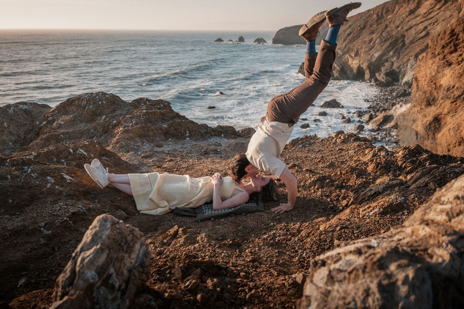 Couple poses playfully on rocky coastal cliffs at sunset, man balancing upside down while woman reclines below.