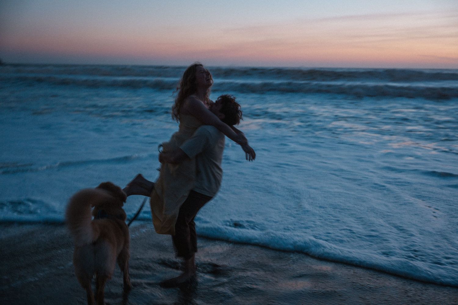 Couple embracing on a beach at sunset with a dog nearby, waves crashing in the background.