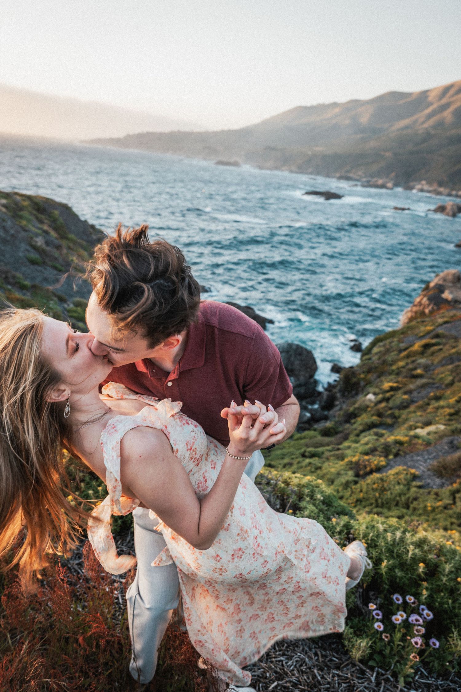 Couple sharing a romantic kiss on a coastal cliff overlooking the ocean at sunset in Big Sur, California.