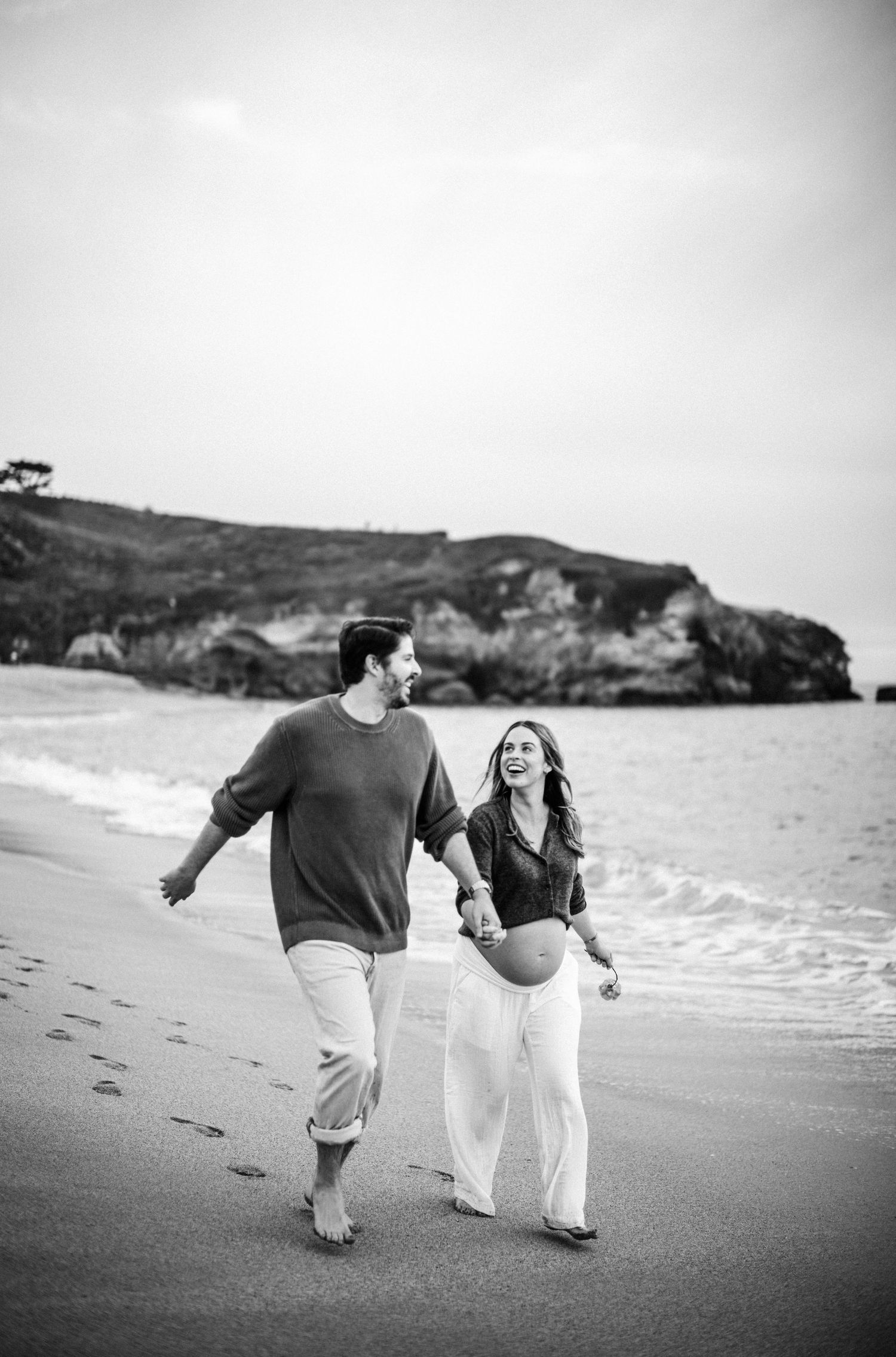 Black and white photo of happy expecting couple walking hand in hand on a beach with rocky cliffs behind them.